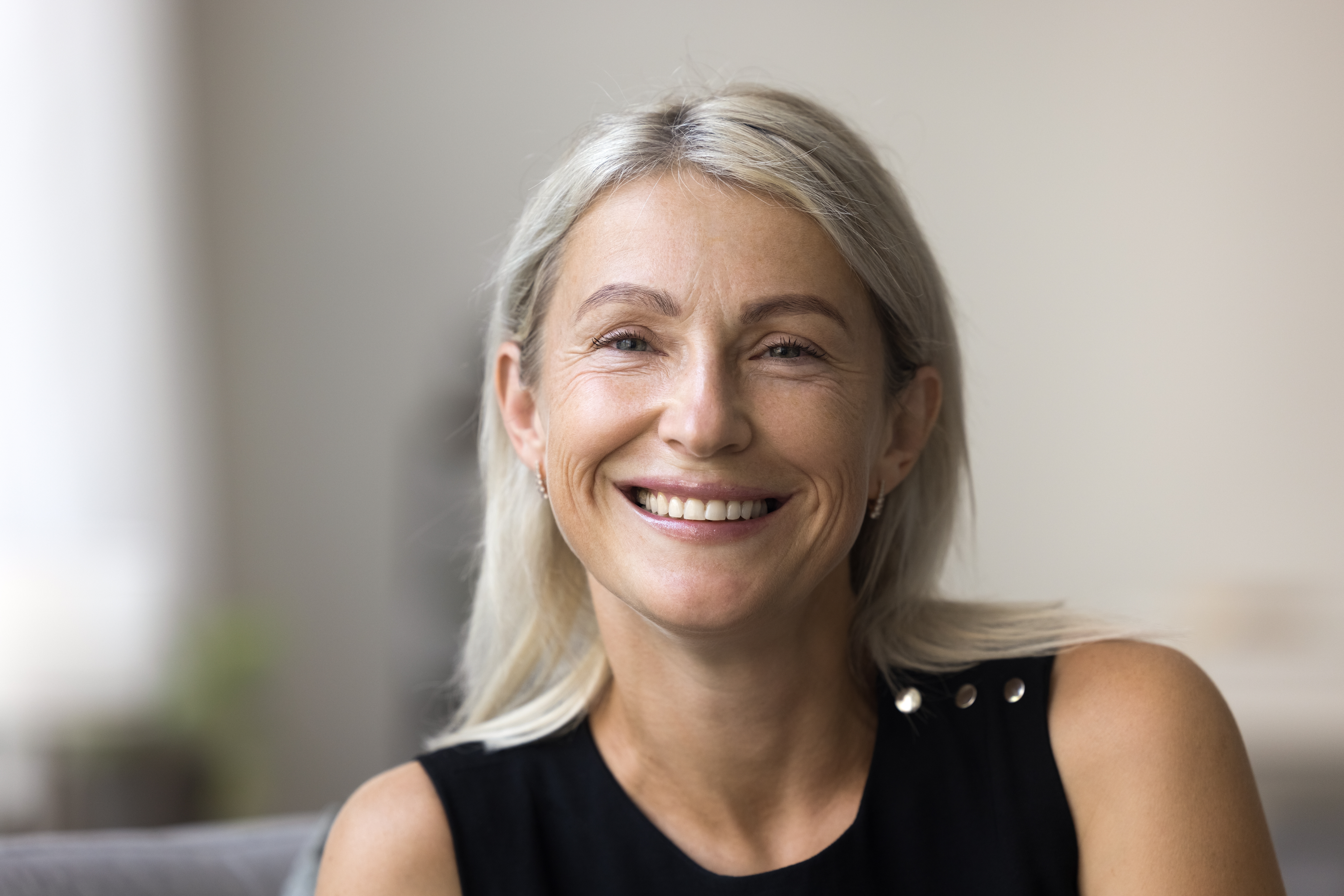 Smiling middle-aged woman with long blonde hair wearing a sleeveless black top, sitting indoors in a softly lit setting
